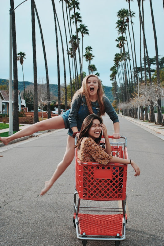 Two young women laughing and having fun with a red shopping cart on a palm tree-lined suburban street. One woman sits inside the cart while the other pushes from behind with an exaggerated expression and one leg extended mid-air.