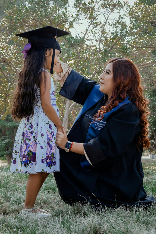 A woman in a graduation gown kneels on the grass, lovingly adjusting the graduation cap of a young girl in a floral dress with long hair and a purple bow. They are holding hands and smiling at each other in a park setting.