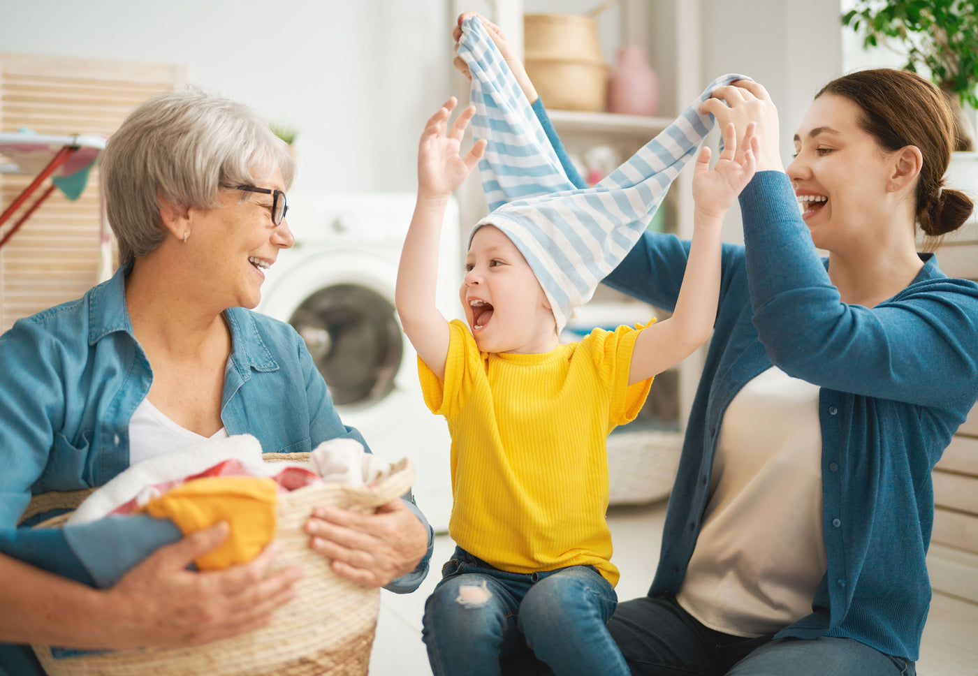 Three generations sharing a joyful moment in the laundry room: a smiling grandmother holds a basket of clothes, while a mother playfully pulls a striped shirt over a laughing child's head.